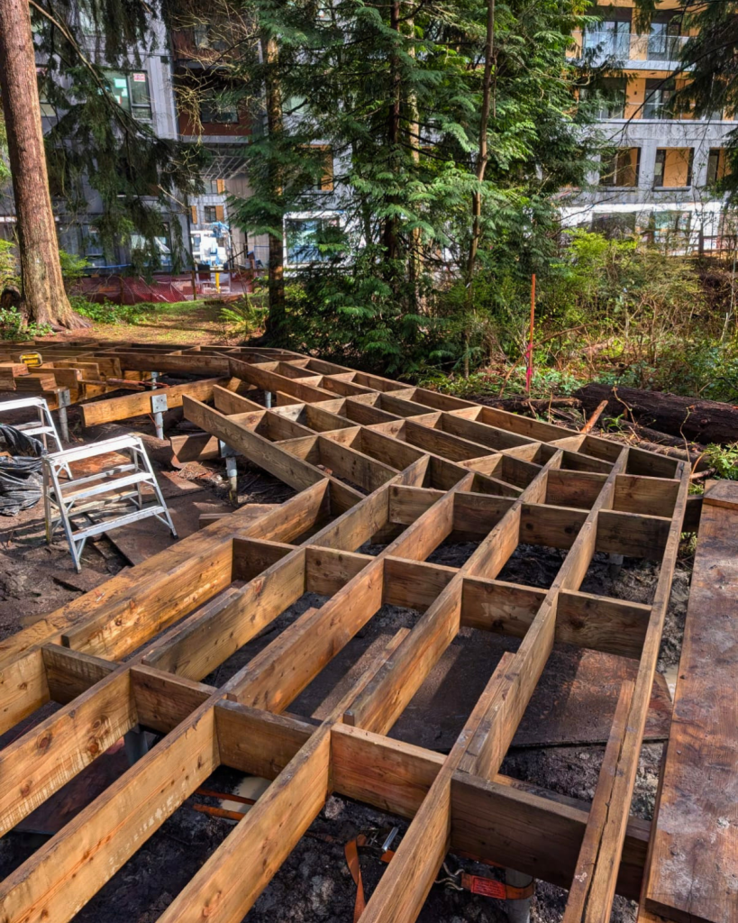 Wooden joist framing installed on helical pile brackets with visible pile caps in a muddy forested construction site