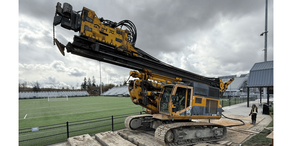 Wide shot of yellow drilling rig on ground protection mats beside the Vancouver FC soccer field under overcast skies