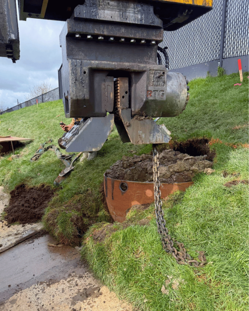 Caisson steel casing installed at ground level beside a grassed slope with a chain and excavated soil visible at the opening