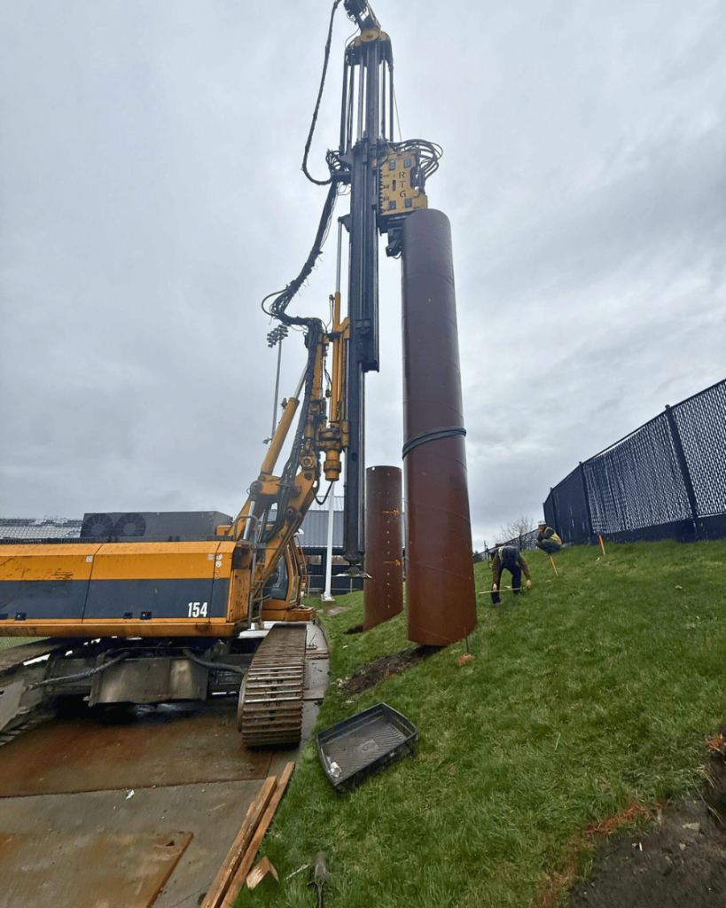Close-up of drilling rig mast and large diameter steel caisson casing with crew members and blue construction fencing visible in background