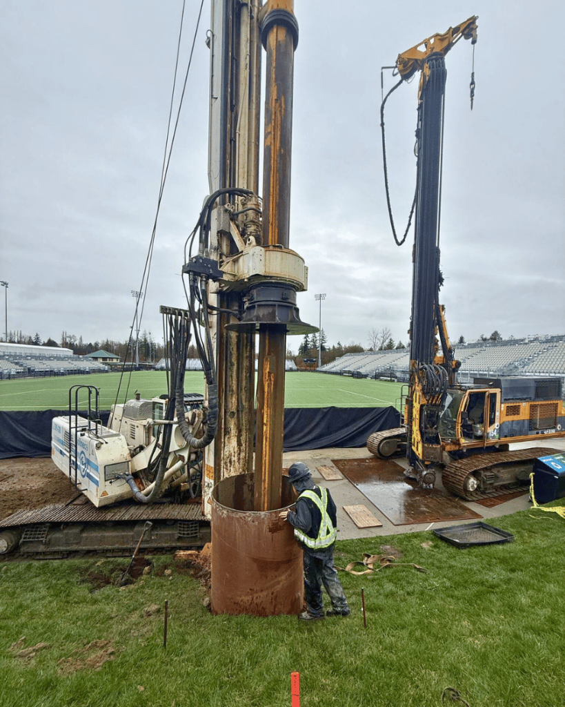TerraCana crew member in high-visibility vest monitoring SoilMec SR30 drill rig augering soil from inside caisson casing on ground protection mats