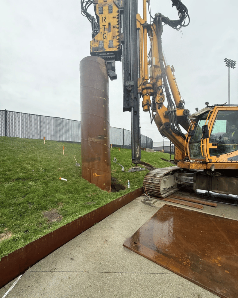Large steel caisson casing installed in the ground with yellow drilling rig positioned alongside on timber ground protection mats, soccer field fence visible in background