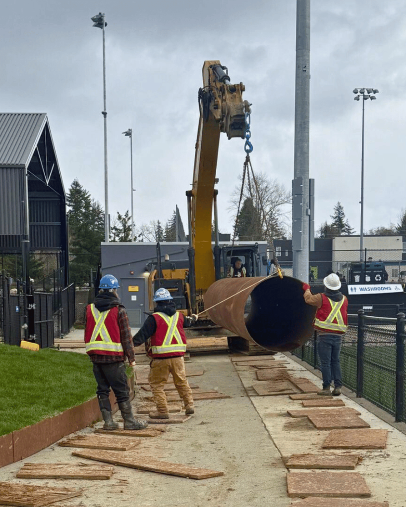 Three workers in high-visibility vests are observing caisson drilling operations beside the Langley Events Centre soccer field