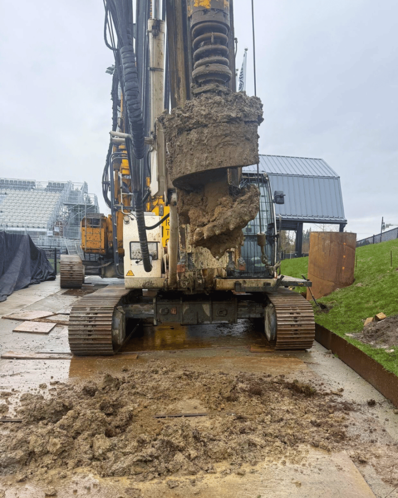 Drilling rig auger removing spoils from inside a caisson casing with excavated soil visible on ground protection mats
