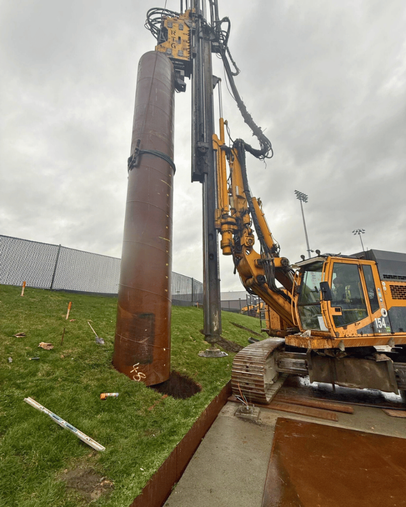 Large diameter steel caisson casing being driven into the ground by a vibratory pile rig adjacent to the Langley Events Centre soccer field