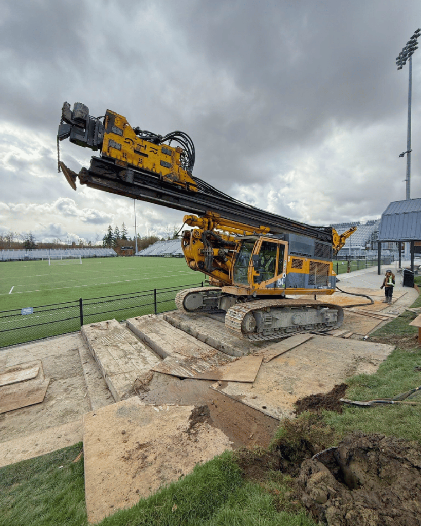 Wide shot of yellow drilling rig on ground protection mats beside the Vancouver FC soccer field under overcast skies